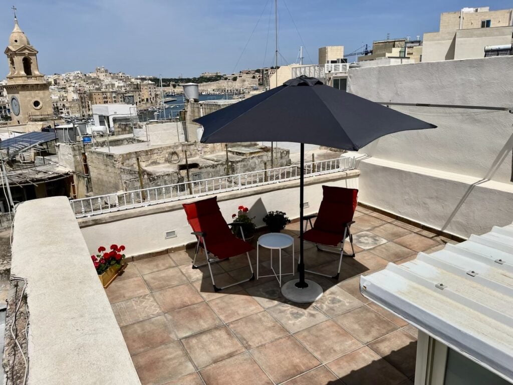 A rooftop terrace with two red chairs and a small round table under a large black umbrella. Potted red flowers are near the railing. The background shows a cityscape with historic buildings and a distant body of water under a clear blue sky.