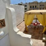 A rooftop terrace with a tiled floor features a shaded canopy area. The canopy has yellow curtains and shelters a white table set with red chairs. Surrounding buildings with traditional architecture are visible in the background under a clear blue sky.