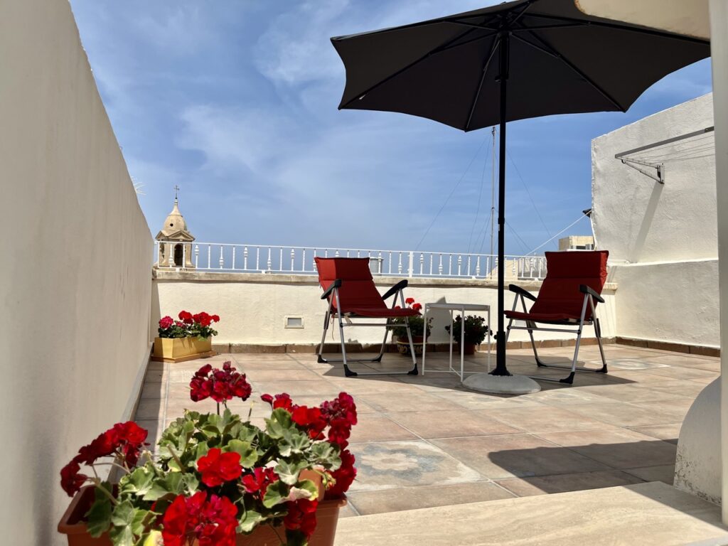 A rooftop patio with two red chairs under a black umbrella. There's a small white table between the chairs. Red flowers in the foreground add a touch of color, with a clear blue sky overhead.