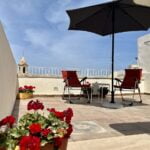 A rooftop patio with two red chairs under a black umbrella. There's a small white table between the chairs. Red flowers in the foreground add a touch of color, with a clear blue sky overhead.