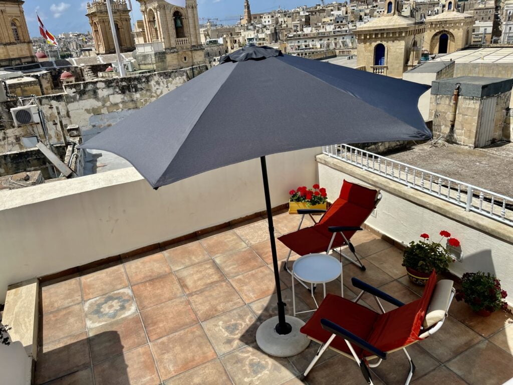 A rooftop patio with two red lounge chairs, a round white table, and a large gray umbrella. The setting overlooks a cityscape with historic buildings under a clear blue sky. Potted flowers add a touch of color.