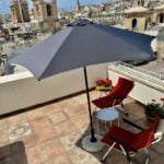 A rooftop patio with two red lounge chairs, a round white table, and a large gray umbrella. The setting overlooks a cityscape with historic buildings under a clear blue sky. Potted flowers add a touch of color.