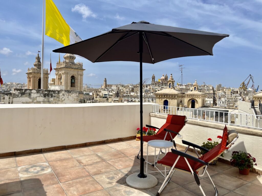 A rooftop terrace with two red folding chairs under a black umbrella overlooks a cityscape with historic church towers. A large yellow and white flag flies in the background. Potted red flowers line the edge of the terrace.