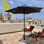 A rooftop terrace with two red folding chairs under a black umbrella overlooks a cityscape with historic church towers. A large yellow and white flag flies in the background. Potted red flowers line the edge of the terrace.