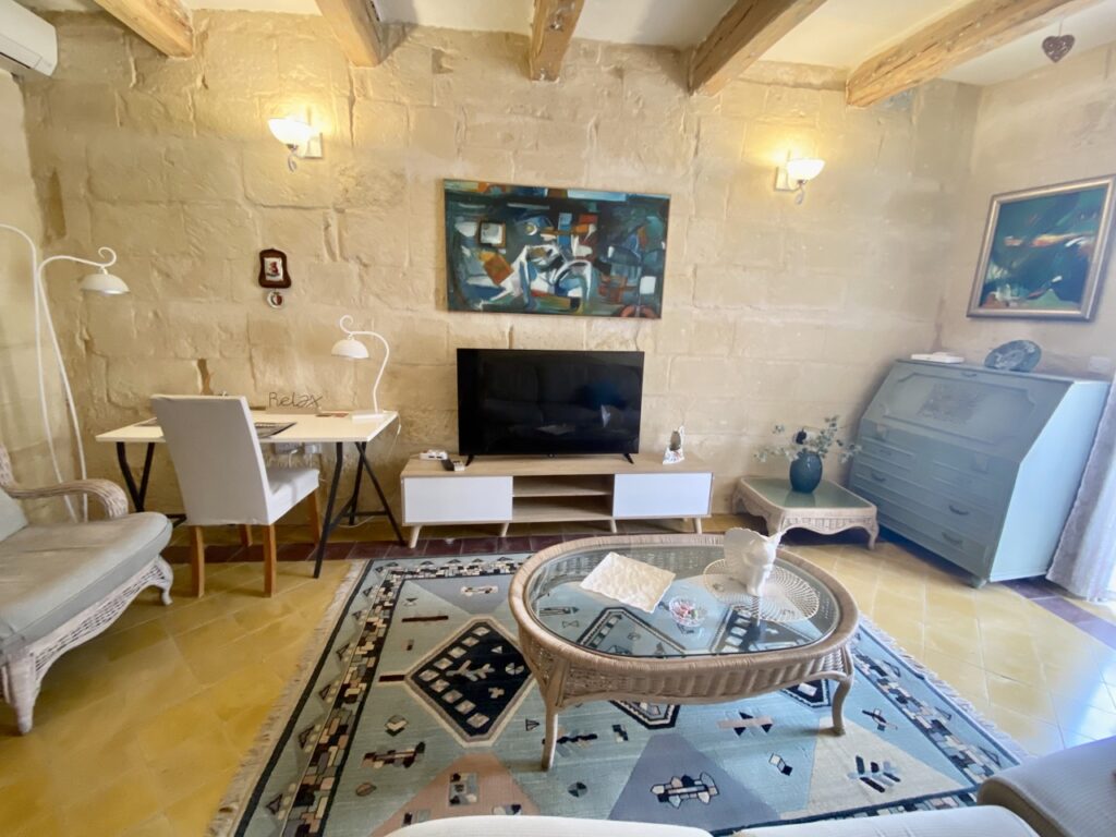 A cozy living room with stone walls features a flat-screen TV on a white stand, a round glass coffee table, and wicker furniture. A desk with a chair sits in the corner, and abstract art hangs on the walls under warm lighting.