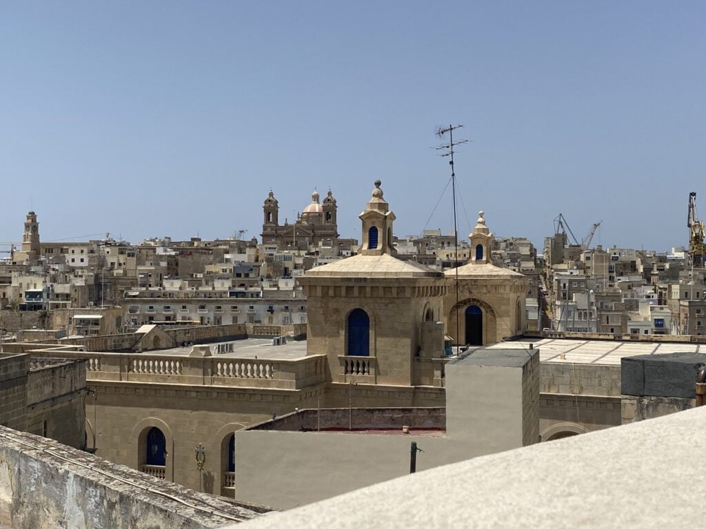 A panoramic view of a city with beige stone buildings under a clear blue sky. Two domed structures with turrets are prominent in the foreground. In the distance, cranes and more buildings are visible.