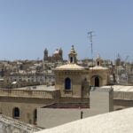 A panoramic view of a city with beige stone buildings under a clear blue sky. Two domed structures with turrets are prominent in the foreground. In the distance, cranes and more buildings are visible.