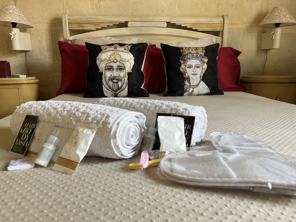 A neatly made bed with red pillows and two decorative portrait pillows. In the foreground are rolled white towels, travel-sized toiletries, and a pair of soft slippers. The bedroom features warm lighting and a cozy ambiance.