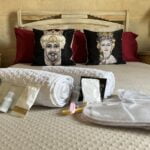 A neatly made bed with red pillows and two decorative portrait pillows. In the foreground are rolled white towels, travel-sized toiletries, and a pair of soft slippers. The bedroom features warm lighting and a cozy ambiance.