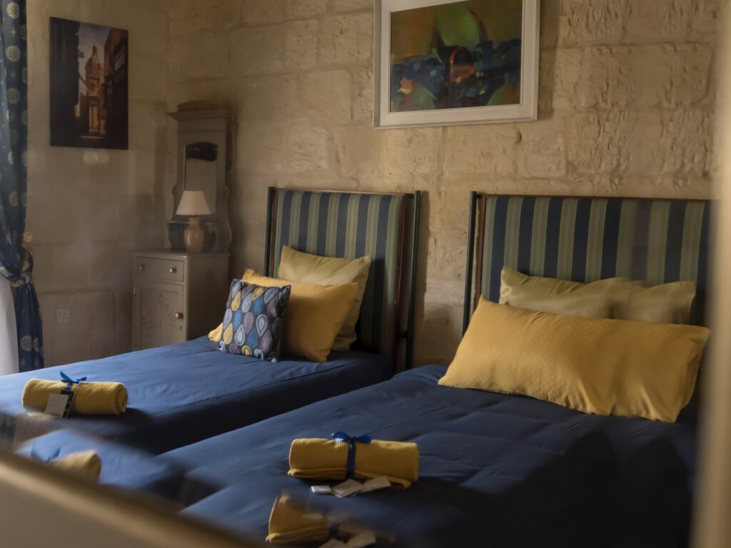 A cozy bedroom with two beds, each adorned with blue and yellow bedding and pillows. Rolled yellow towels tied with blue ribbons sit at the foot of the beds. A lamp on a nightstand and a painting on the wall add warmth to the stone room.