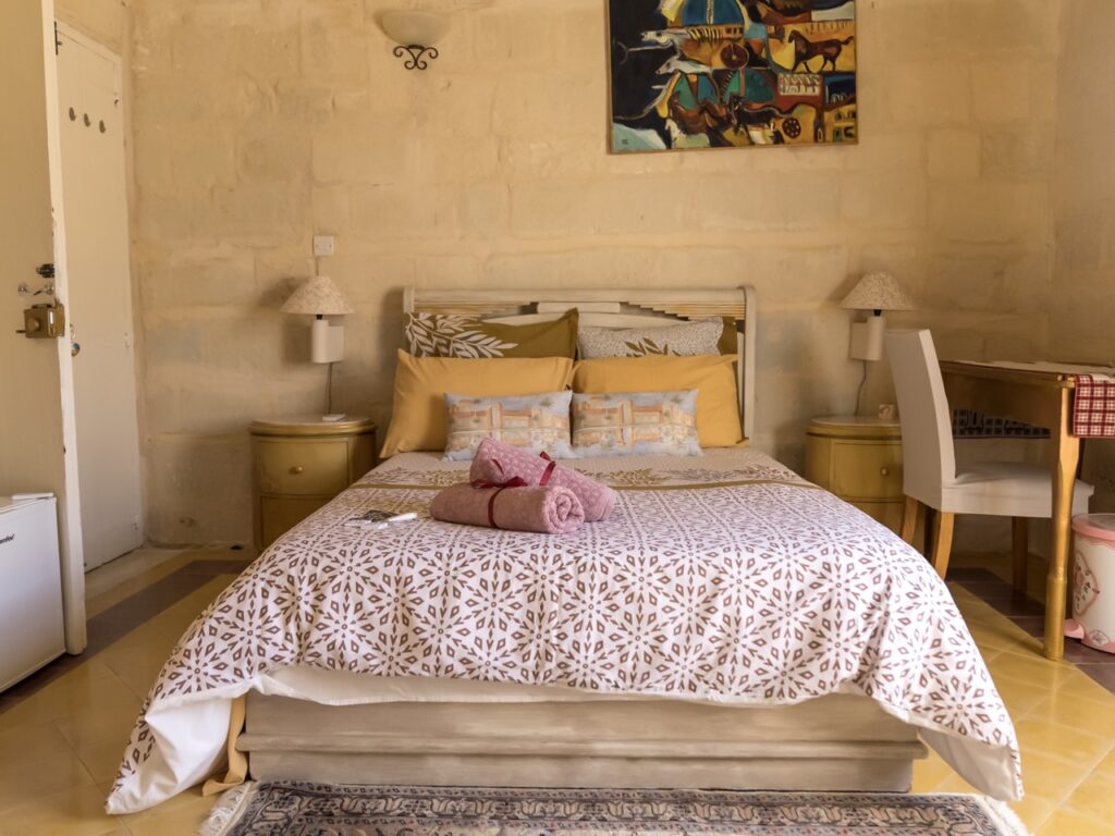 A cozy bedroom with a double bed featuring patterned bedding and rolled towels. The room includes two nightstands with lamps, a desk with a chair, and a painting on the textured wall. Natural light enhances the warm, inviting ambiance.