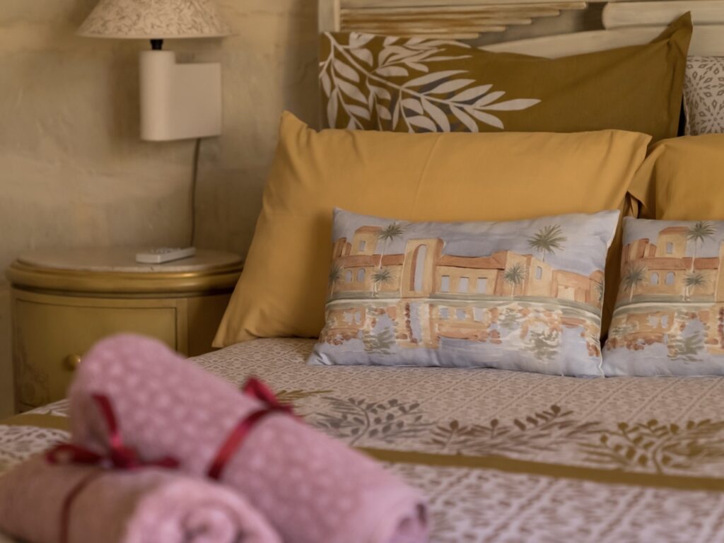 A neatly made bed with yellow and patterned pillows, featuring a decorative pillow with an illustration of a building and trees. Two rolled pink towels are placed at the foot of the bed. A small lamp and a round nightstand are beside the bed.
