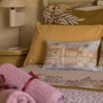 A neatly made bed with yellow and patterned pillows, featuring a decorative pillow with an illustration of a building and trees. Two rolled pink towels are placed at the foot of the bed. A small lamp and a round nightstand are beside the bed.