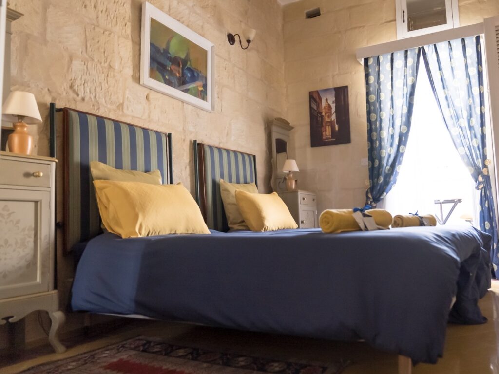A cozy bedroom with two striped beds, blue bedding, and yellow pillows. Light streams through a window with patterned curtains. The stone walls display framed artwork, and lamps sit on elegant bedside tables. Rolled towels are placed on the beds.