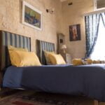 A cozy bedroom with two striped beds, blue bedding, and yellow pillows. Light streams through a window with patterned curtains. The stone walls display framed artwork, and lamps sit on elegant bedside tables. Rolled towels are placed on the beds.