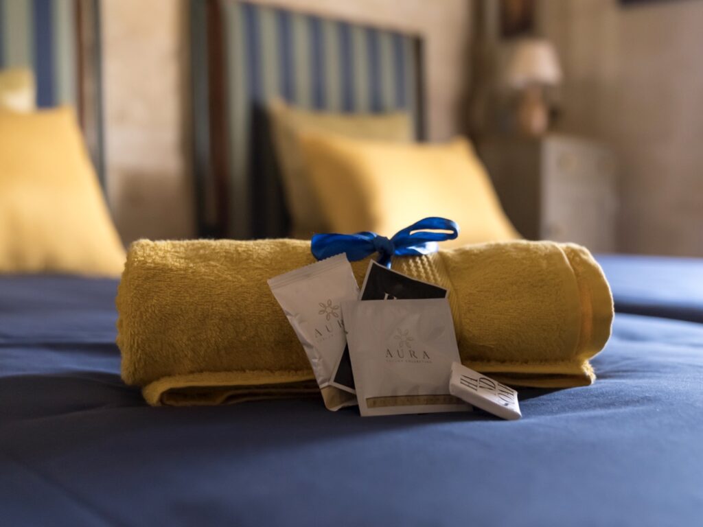 A yellow towel tied with a blue ribbon is placed on a blue bedspread. Beside it, there are packets labeled "Aura." The background shows a blurred bedroom with striped headboards and a nightstand.