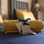 A yellow towel tied with a blue ribbon is placed on a blue bedspread. Beside it, there are packets labeled "Aura." The background shows a blurred bedroom with striped headboards and a nightstand.