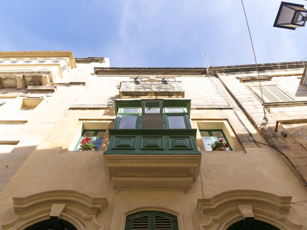 View from below of a traditional building facade with a green enclosed balcony and potted plants. Bright blue sky above, with part of a streetlamp and nearby structures visible.