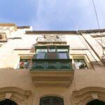 View from below of a traditional building facade with a green enclosed balcony and potted plants. Bright blue sky above, with part of a streetlamp and nearby structures visible.