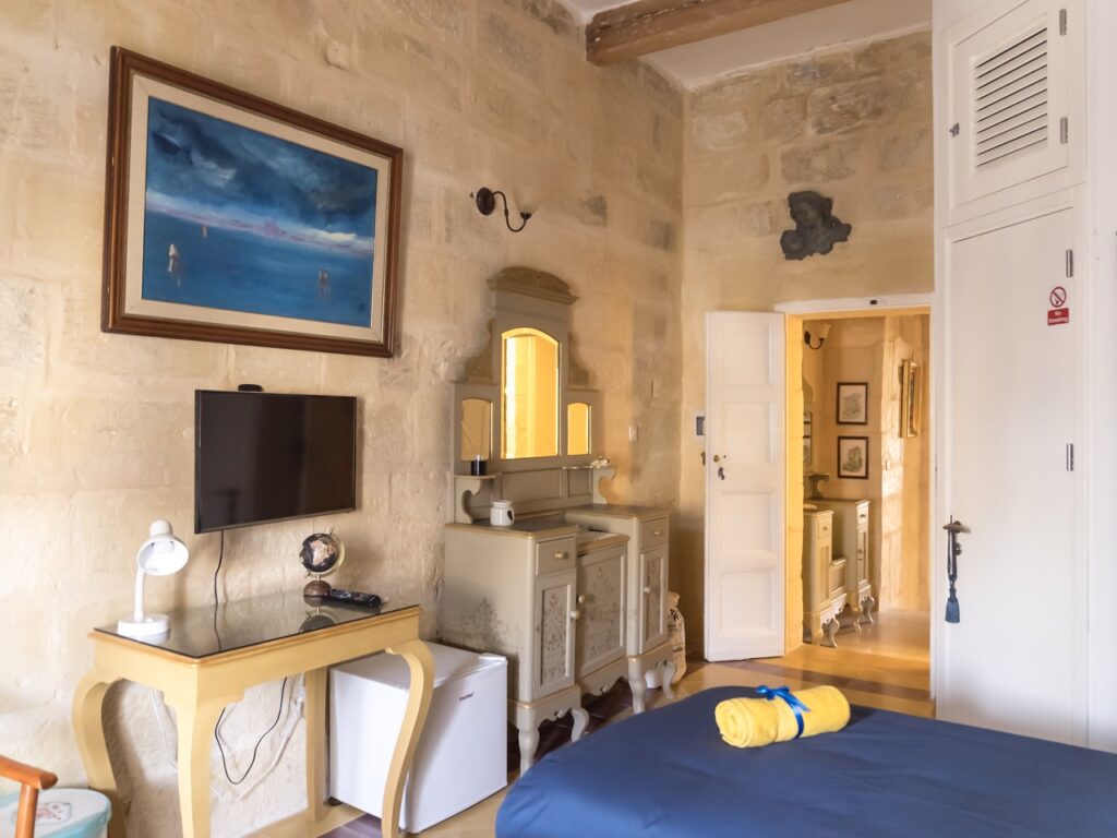 A cozy room with stone walls features a bed with a blue bedspread, a wall-mounted TV, a wooden vanity with a mirror, and a mini fridge. An abstract painting is hung on the wall above the TV. An open door reveals a small corridor.