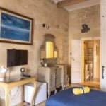 A cozy room with stone walls features a bed with a blue bedspread, a wall-mounted TV, a wooden vanity with a mirror, and a mini fridge. An abstract painting is hung on the wall above the TV. An open door reveals a small corridor.