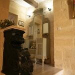 A cozy hallway with stone walls and soft lighting. A decorative dresser with a large mirror is adorned with a teddy bear. An ornate black banister is visible in the foreground, and framed pictures hang on the walls.
