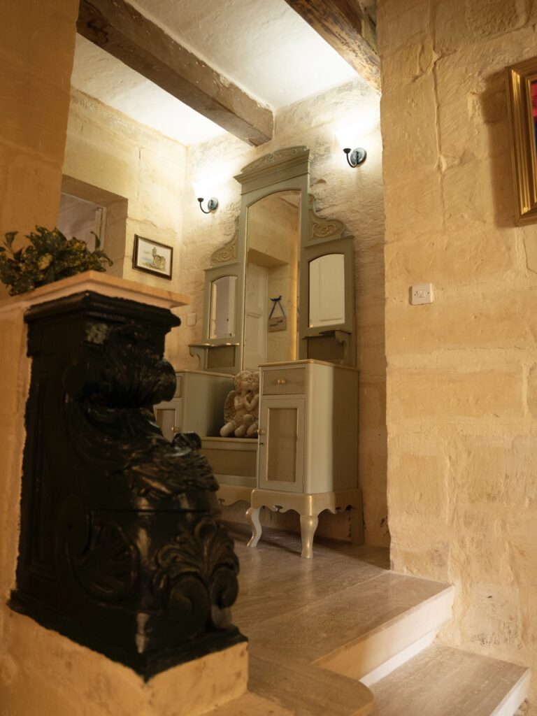 A cozy hallway with stone walls and soft lighting. A decorative dresser with a large mirror is adorned with a teddy bear. An ornate black banister is visible in the foreground, and framed pictures hang on the walls.