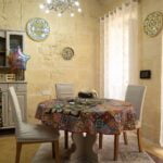 A cozy dining room with a round table covered in a colorful tablecloth. The table is set for two, and a birthday balloon is attached to one chair. Decorative plates adorn the pale stone walls. A modern chandelier hangs overhead, and soft curtains cover a window.