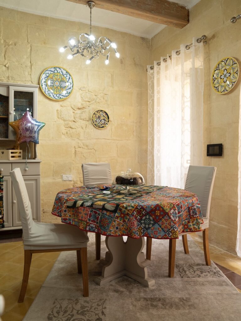 A cozy dining room with a round table covered in a colorful tablecloth. The table is set for two, and a birthday balloon is attached to one chair. Decorative plates adorn the pale stone walls. A modern chandelier hangs overhead, and soft curtains cover a window.