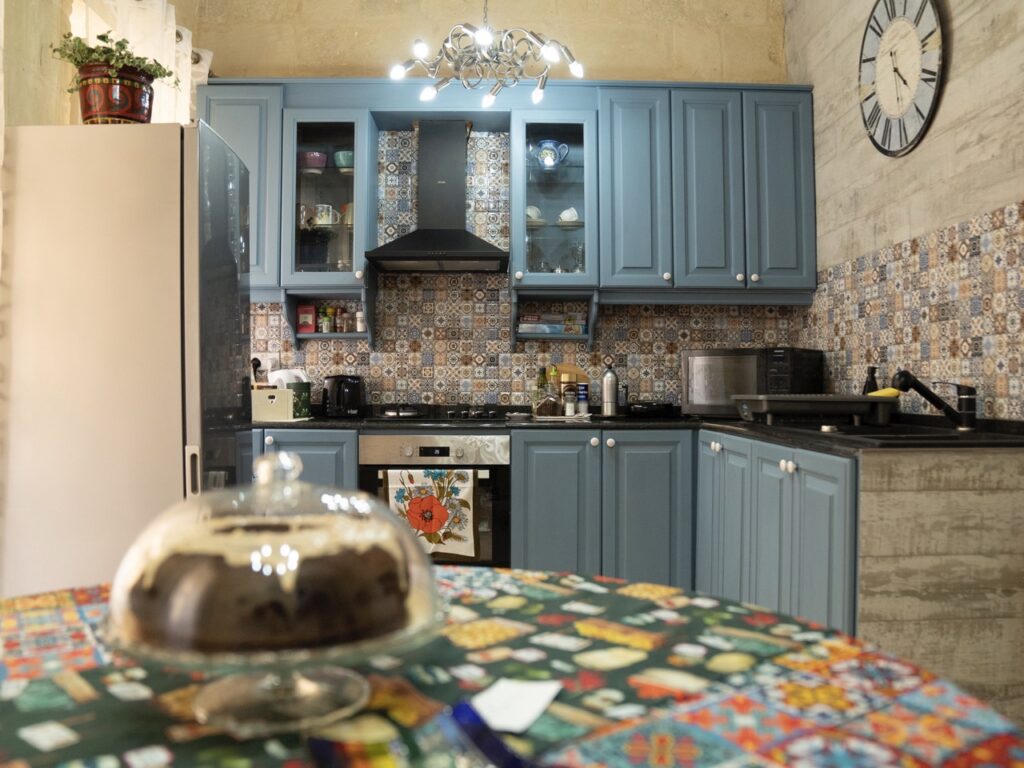 A cozy kitchen with blue cabinets and decorative tile backsplash. A round table in the foreground has a colorful tablecloth and a glass cake stand. A large clock is on the wall, and modern appliances fill the space.