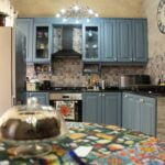 A cozy kitchen with blue cabinets and decorative tile backsplash. A round table in the foreground has a colorful tablecloth and a glass cake stand. A large clock is on the wall, and modern appliances fill the space.