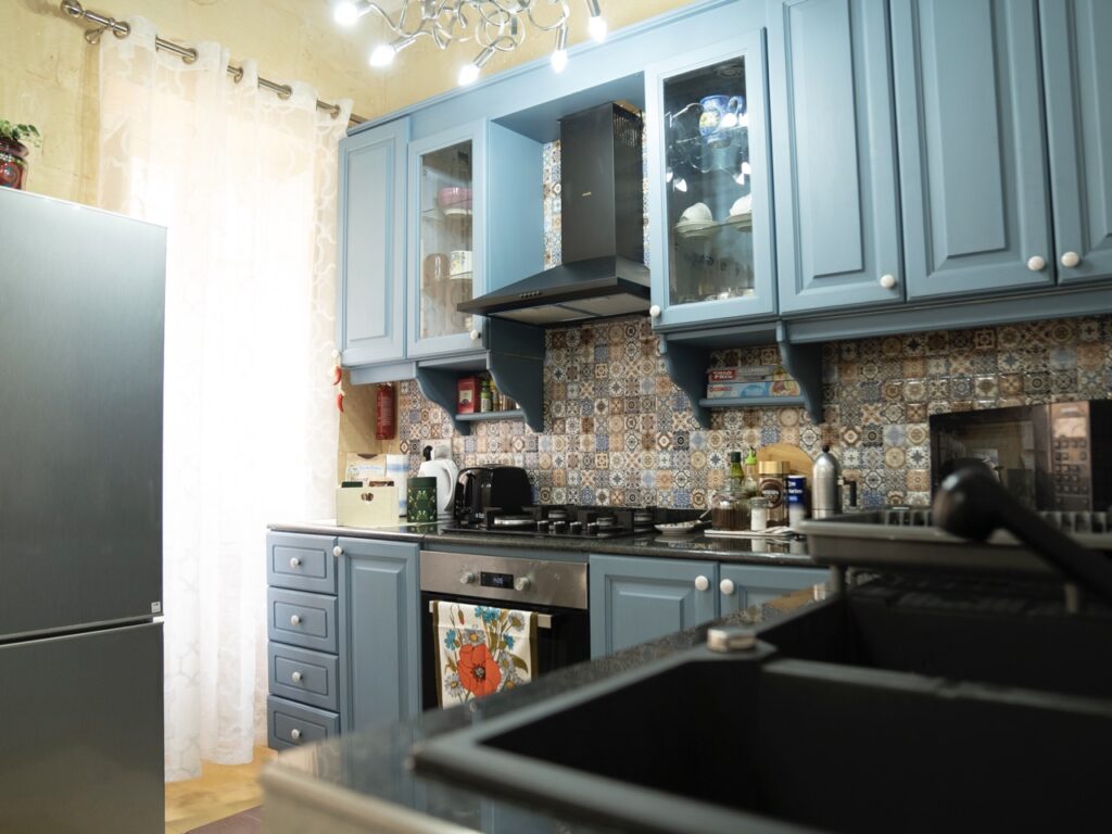 A cozy kitchen with blue cabinets and a patterned backsplash. There is a stainless steel refrigerator, a stove with a black range hood, and various small appliances on the counter. Soft lighting and sheer curtains add to the warm atmosphere.