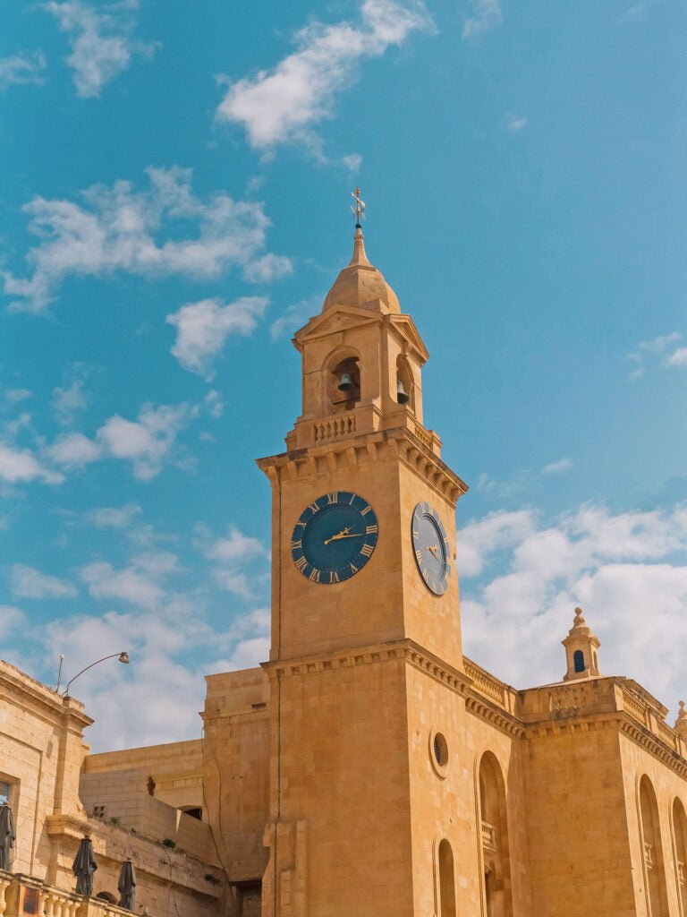 A tall, beige stone clock tower with a pointed roof stands against a bright blue sky with scattered clouds, reminiscent of the classical architecture often seen in Malta vacation spots. The clock face is blue with gold numbers, adding to its timeless appeal.