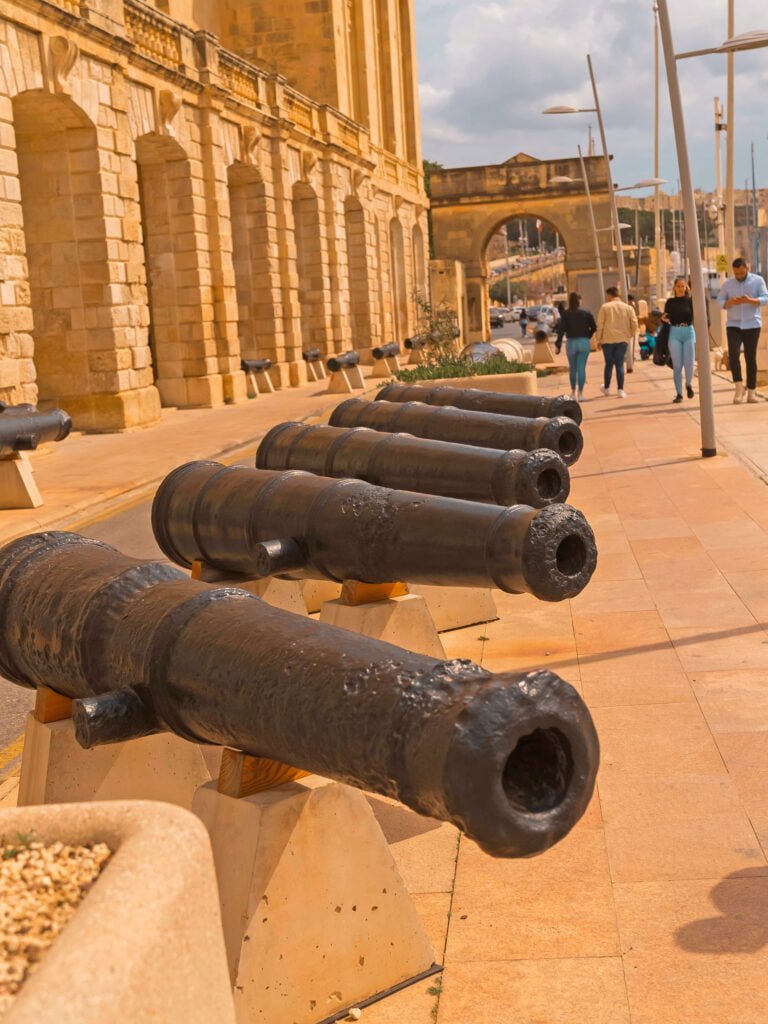A row of old black cannons displayed on a stone pathway lined with historic buildings in Malta sets the scene for an unforgettable vacation. People walk nearby under a clear blue sky, with an arched stone entrance visible in the background.