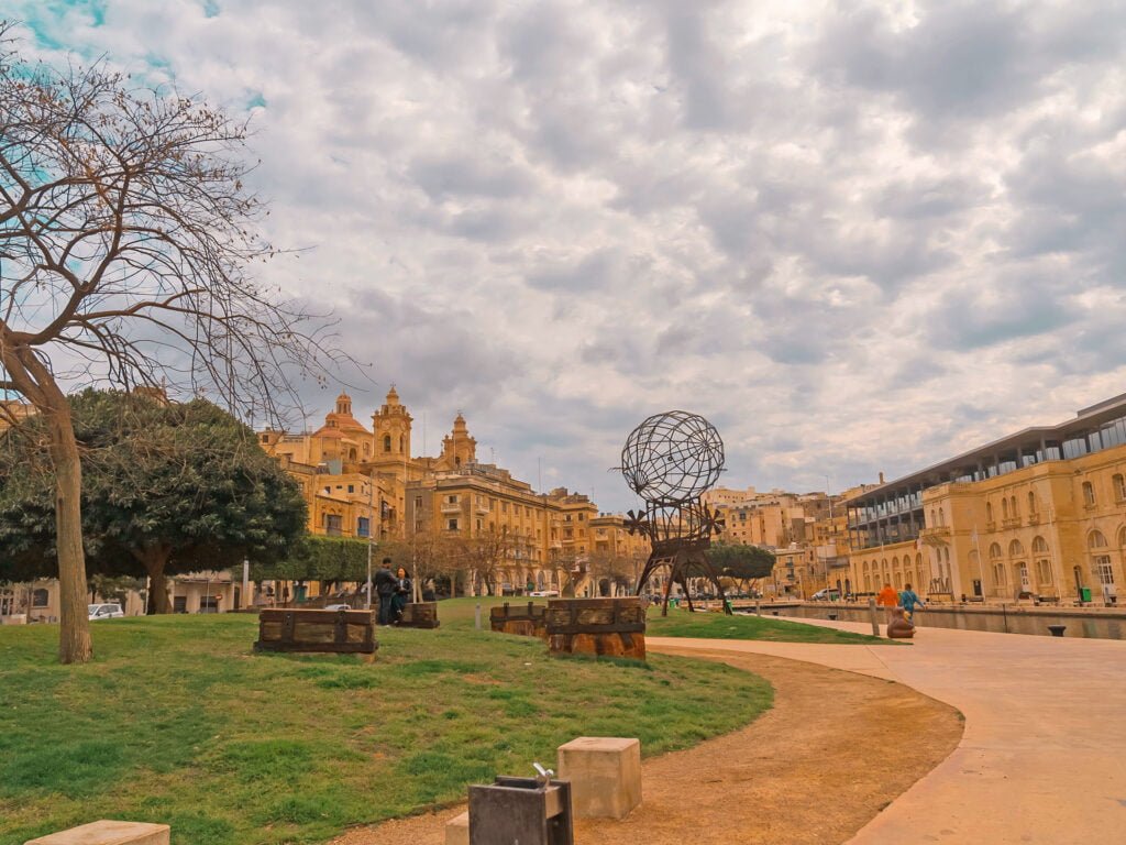 A scenic park in Malta features a path leading to a globe-shaped sculpture. Historic stone buildings and church towers rise under the cloudy sky, evoking classic Malta holidays vibes. Bare trees and benches are scattered across the grassy area, offering a serene escape for your Malta vacation.