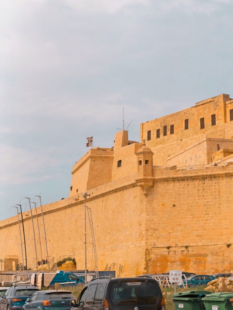 A stone fortress with high walls and multiple towers stands under a partly cloudy sky. Several vehicles and sailboats are parked in the foreground near a line of green dumpsters, evoking a historic, coastal atmosphere typical of Malta holidays.