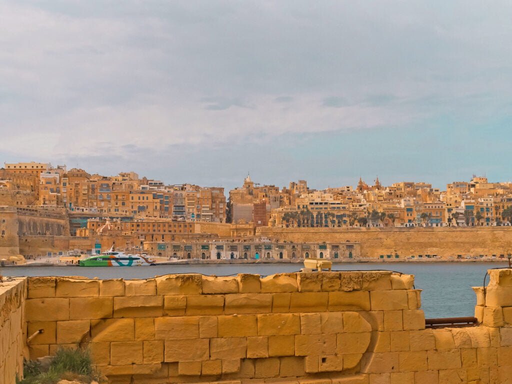 View of a historic coastal city in Malta, with warm-toned buildings along the water's edge. In the foreground, a stone wall is visible. The sky is overcast, casting a soft light over this picturesque scene, perfect for those seeking relaxing Malta holiday homes.