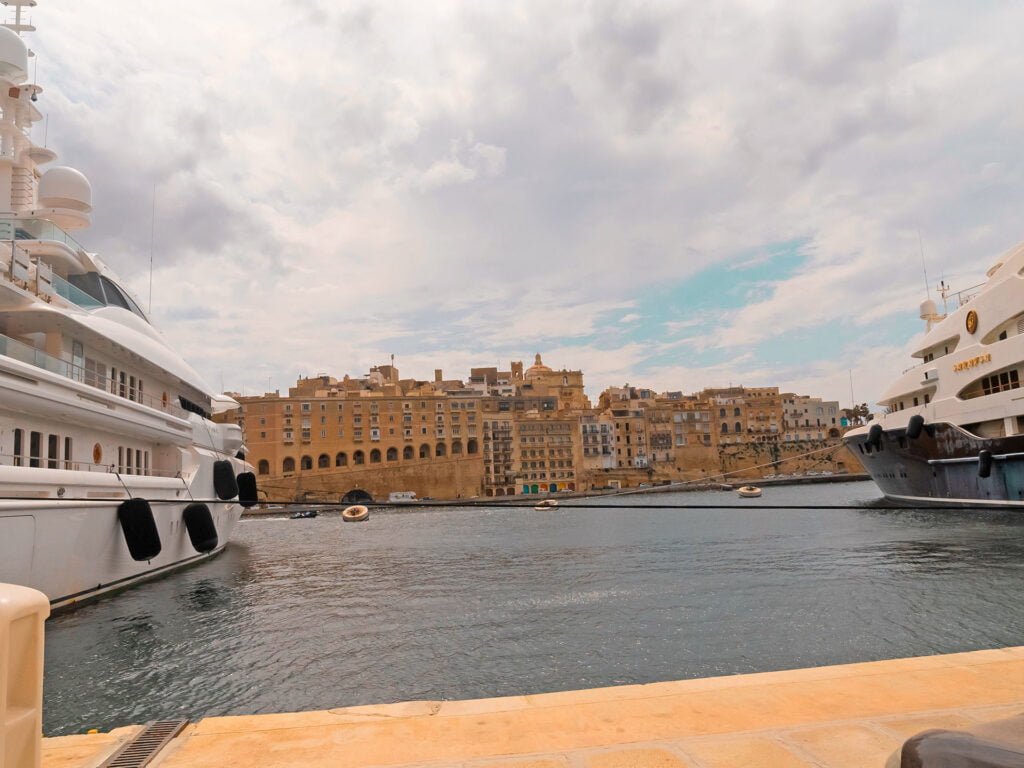 A waterfront scene with two large yachts docked on either side. Behind them is a historic cityscape, reminiscent of Malta holiday homes, with beige stone buildings under a cloudy sky reflecting on the calm water.