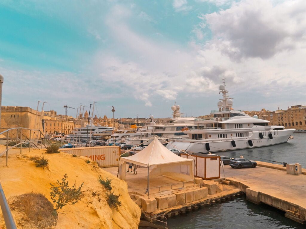 A harbor scene with multiple large yachts docked along the water. Beige buildings resembling Malta holiday homes and cranes are visible in the background under a partly cloudy sky. A small tent and greenery are in the foreground on a stone platform.